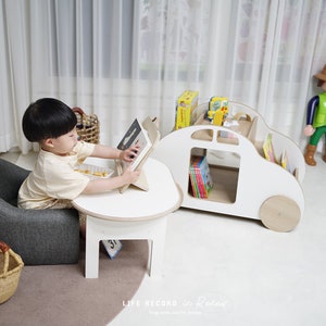 May include: A young child sits in a gray armchair and uses a tablet at a white table. A white wooden toy car with shelves for books is in the background. The car has a brown steering wheel and brown wheels.