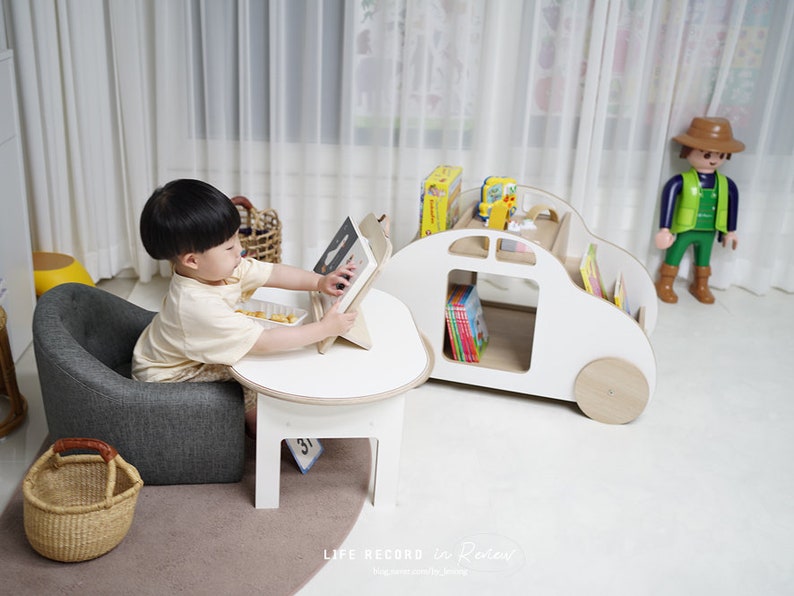 May include: A young child sits in a gray armchair and uses a tablet at a white table. A white wooden toy car with shelves for books is in the background. The car has a brown steering wheel and brown wheels.