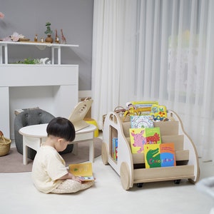 May include: A white wooden toy car with wheels, used as a bookshelf, is filled with colorful children's books. A young child sits on the floor reading a book.