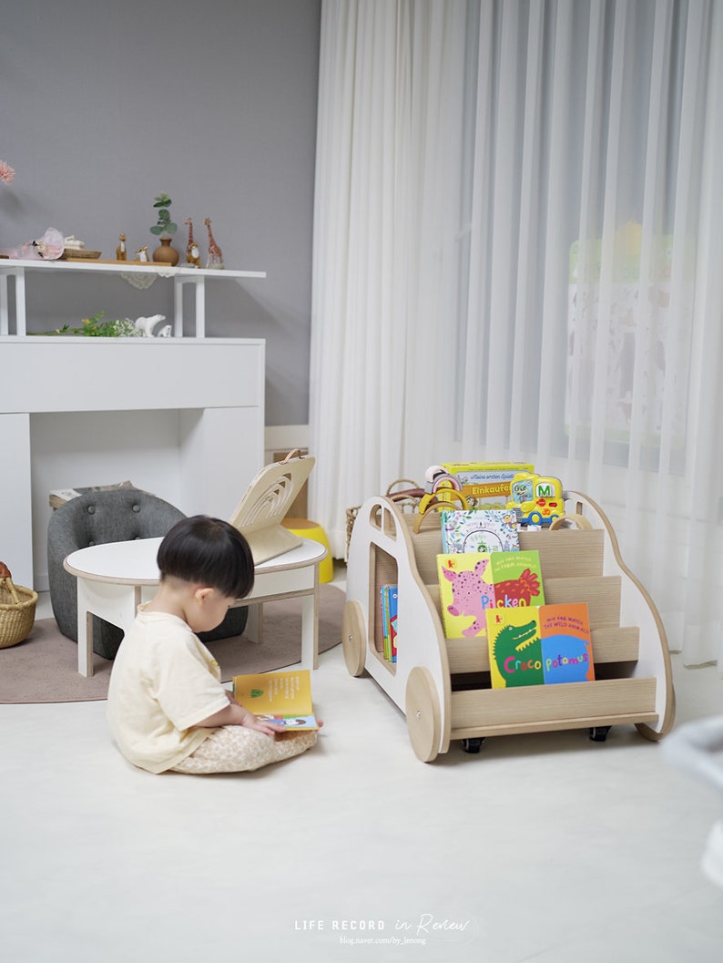May include: A white wooden toy car with wheels, used as a bookshelf, is filled with colorful children's books. A young child sits on the floor reading a book.