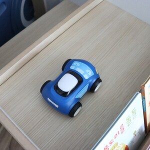 May include: A blue toy car with white details sits on a wooden shelf. The car is on a shelf with books behind it.