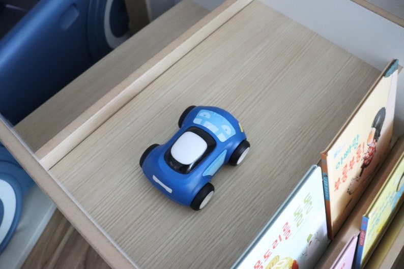 May include: A blue toy car with white details sits on a wooden shelf. The car is on a shelf with books behind it.