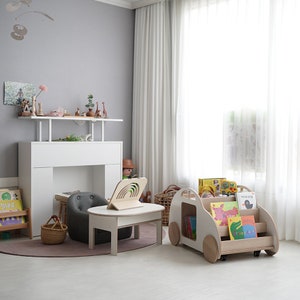 May include: A white wooden toy car with wheels, used as a bookshelf, filled with colorful children's books. The car is on a light wood floor in a room with a white table and a gray chair.