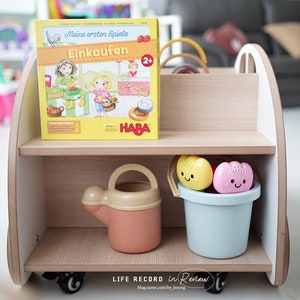 May include: A wooden toy shelf with two shelves. The top shelf has a German board game box with the title "Meine ersten Spiele Einkaufen" and a HABA logo. The bottom shelf has a blue bucket with two colorful bath toys and a yellow watering can.
