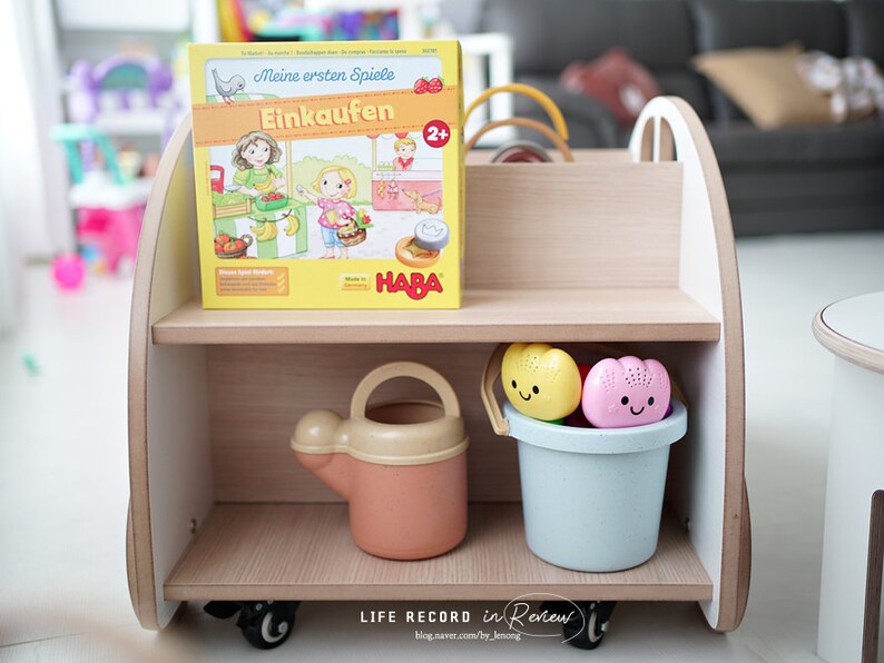May include: A wooden toy shelf with two shelves. The top shelf has a German board game box with the title "Meine ersten Spiele Einkaufen" and a HABA logo. The bottom shelf has a blue bucket with two colorful bath toys and a yellow watering can.