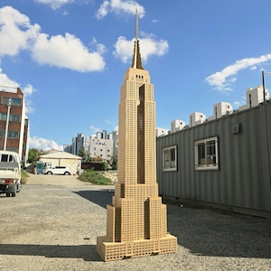 Puede incluir: Una maqueta detallada del Empire State Building, construida con materiales de color marrón claro. El modelo reproduce el diseño icónico del edificio, incluyendo su aguja y numerosas ventanas. El modelo se alza sobre un fondo de cielo azul y edificios circundantes.
