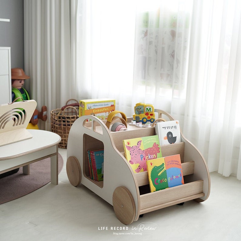 May include: A white wooden toy car with wheels, shelves, and a storage compartment. The car is filled with colorful children's books and toys. The car is on a white floor in a room with white curtains.