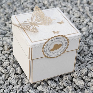 May include: A white square box with a gold trim and a gold butterfly design. The box has a white lace trim and a gold sticker with the text "Congratulations" on it.