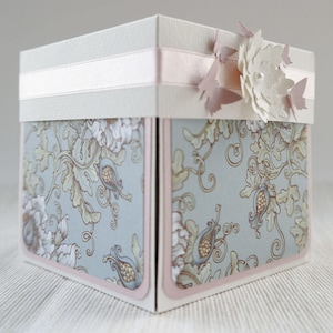 Könnte beinhalten: Eine weiße Schachtel mit einem hellrosa Band und floralen Verzierungen. Die Schachtel ist mit einem blauen und weißen Blumenmuster verziert.