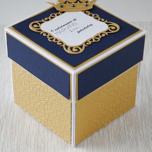 May include: A gold and blue box with a white top. The top has a gold crown and a white label with the text "I anledning av MARVINS 1 års fødselsdag".