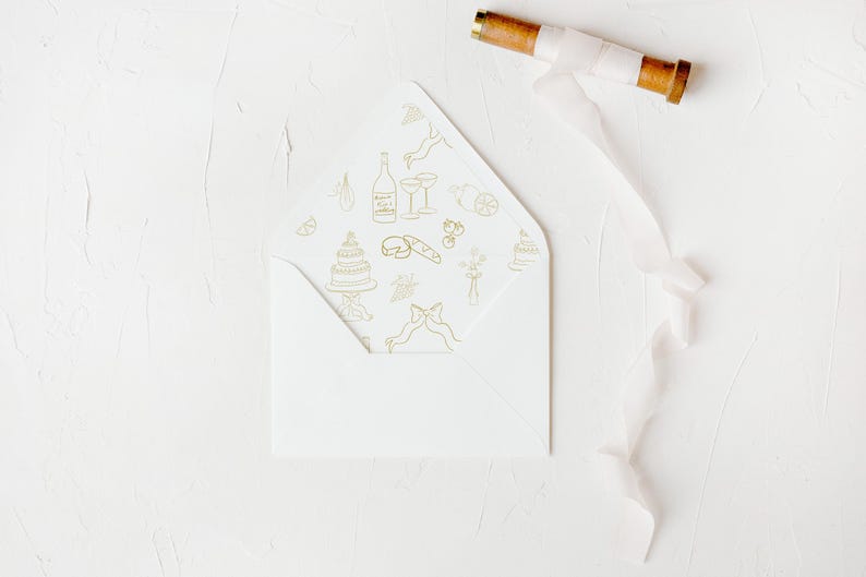 May include: A white envelope with a gold pattern of wedding-related icons, including a cake, champagne glasses, and a bottle of champagne. The envelope is lying on a white surface with a white ribbon.