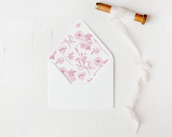 May include: A white envelope with a pink floral pattern on the flap. The envelope is lying on a white textured surface with a spool of white ribbon.