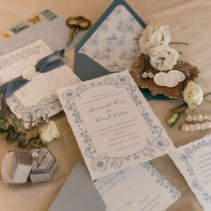 May include: Wedding invitation suite with blue floral designs and white text. The invitation reads "With great joy, we are united to the wedding of Savannah Rose and Raul Vater. Saturday, the thirtieth of May, two thousand twenty two. Two hundred twenty two East Oak Street, Shreveport, Louisiana. Reception to follow."