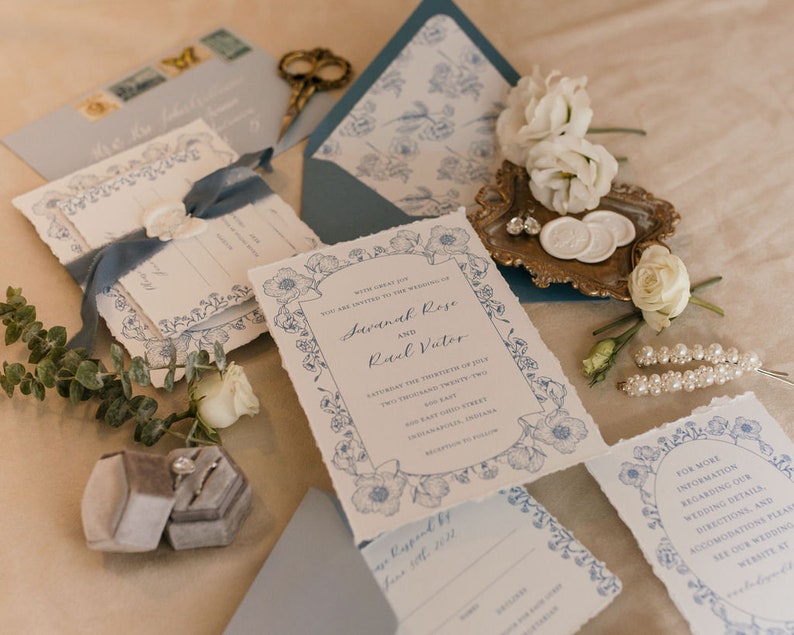 May include: Wedding invitation suite with blue floral designs and white text. The invitation reads "With great joy, we are united to the wedding of Savannah Rose and Raul Vater. Saturday, the thirtieth of May, two thousand twenty two. Two hundred twenty two East Oak Street, Shreveport, Louisiana. Reception to follow."