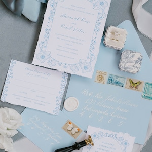 May include: A wedding invitation suite with a blue and white floral design. The invitation card features the names Savanah Rose and Rael Victor, the date Saturday the thirtieth of July, two thousand twenty-two, and the location 600 East Ohio Street, Indianapolis, Indiana. The RSVP card has a floral border and includes the text "Please Respond by June 10th, 2022". The envelope is light blue with white calligraphy and a wax seal.