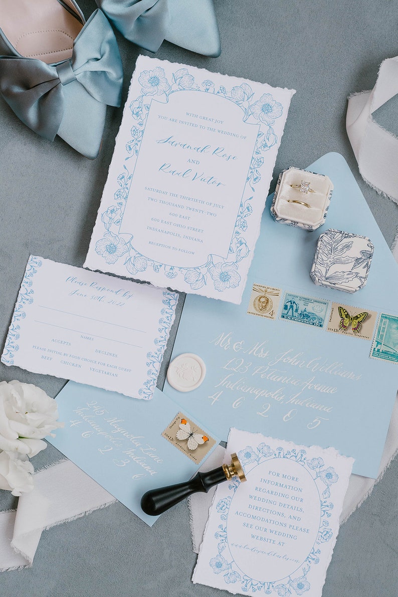 May include: A wedding invitation suite with a blue and white floral design. The invitation card features the names Savanah Rose and Rael Victor, the date Saturday the thirtieth of July, two thousand twenty-two, and the location 600 East Ohio Street, Indianapolis, Indiana. The RSVP card has a floral border and includes the text "Please Respond by June 10th, 2022". The envelope is light blue with white calligraphy and a wax seal.