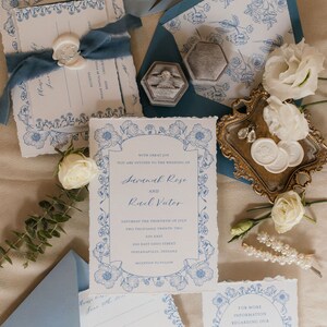 May include: Wedding invitation suite with blue and white floral designs. The invitation card features the names Savannah Rose and Raal Vutor, the date Saturday the thirtieth of July, two thousand twenty-two, and the address 600 East Ohio Street, Indianapolis, Indiana. The invitation is surrounded by a blue and white floral border.