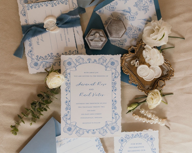May include: Wedding invitation suite with blue and white floral designs. The invitation card features the names Savannah Rose and Raal Vutor, the date Saturday the thirtieth of July, two thousand twenty-two, and the address 600 East Ohio Street, Indianapolis, Indiana. The invitation is surrounded by a blue and white floral border.