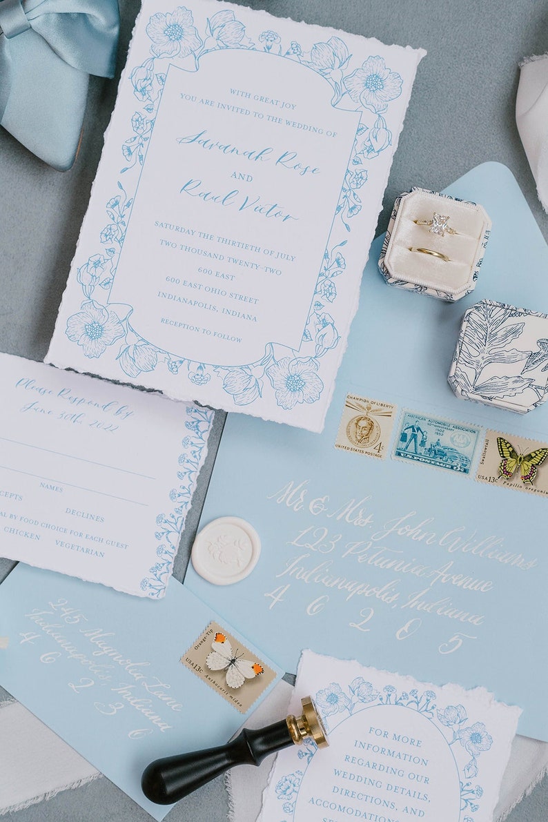 May include: Wedding invitation suite with blue floral designs. The invitation card features the names "Savannah Rose" and "Ravel Victor" with the date "Saturday the Thirtieth of July Two Thousand Twenty-Two". The RSVP card has a blue floral border and the text "Please Respond by June 30th 2022". The envelope is light blue with white calligraphy and a wax seal.