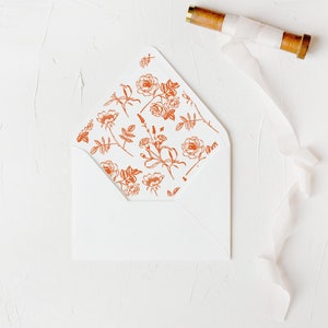 May include: A white envelope with a floral pattern on the flap. The pattern is in a rust orange colour and features small flowers and leaves. The envelope is lying flat on a white surface with a spool of white ribbon.