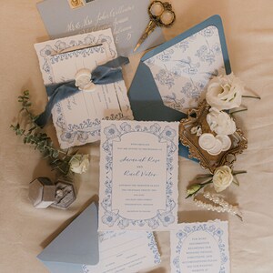 May include: Blue and white floral wedding invitation suite with a wax seal, a ring box, and a small bouquet of white flowers. The invitation includes the names Savanah Rose and Raul Vutor, the date Saturday the thirtieth of July, two thousand twenty-two, and the location 600 East Ohio Street, Indianapolis, Indiana.