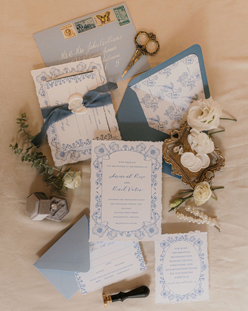 May include: Blue and white floral wedding invitation suite with a wax seal, a ring box, and a small bouquet of white flowers. The invitation includes the names Savanah Rose and Raul Vutor, the date Saturday the thirtieth of July, two thousand twenty-two, and the location 600 East Ohio Street, Indianapolis, Indiana.