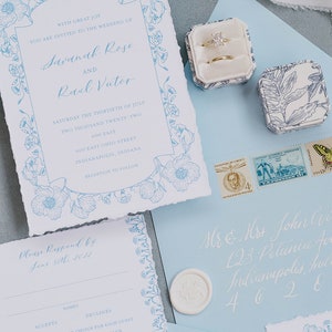 May include: A wedding invitation with a blue floral border and the text "With great joy you are invited to the wedding of Savanah Rose and Rael Victor" on a white background. The invitation includes the date, time, and location of the wedding. The invitation also includes a RSVP card with a blue floral border and the text "Please Respond by June 30th, 2022".