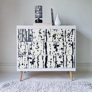 May include: A white three-drawer dresser with a black and white abstract design on the drawer fronts. The dresser has wooden legs and a small lamp, a book, and a white vase on top. A gray rug is in front of the dresser.