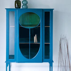 Puede incluir: Un armario azul turquesa con puertas de cristal y estantes. El armario tiene un diseño ovalado con una inserción verde acanalada. Dos libros pequeños están en un estante en el interior. Dos jarrones de cristal y plantas secas encima.