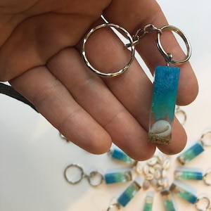 May include: A blue and green resin keychain with a seashell inside. The keychain is attached to a silver key ring. There are several other keychains in the background.