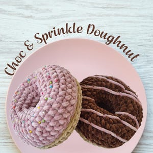 Puede incluir: Dos donuts de ganchillo hechos a mano sobre un plato rosa. Uno es rosa claro con chispas de colores, el otro es marrón con rayas de glaseado rosa. El texto "Choc & Sprinkle Doughnut" y "Crochet Pattern" son visibles.