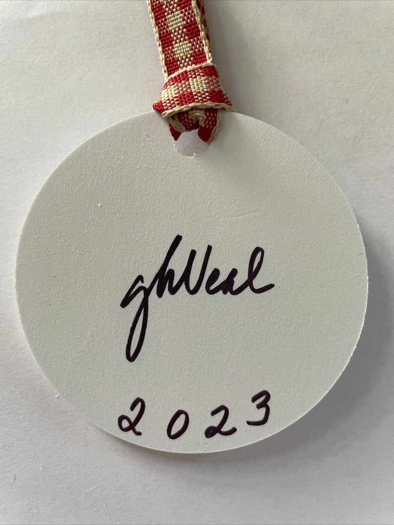 Puede incluir: Adorno redondo blanco con texto manuscrito negro "goal" y el a&ntilde;o "2023" en negro debajo. El adorno est&aacute; sujeto a una cinta de cuadros rojos y blancos.