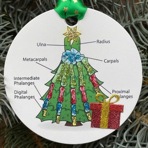 Puede incluir: Un adorno redondo blanco con forma de &aacute;rbol de Navidad con una caja de regalo roja y dorada en la parte inferior. El &aacute;rbol est&aacute; decorado con purpurina y tiene los huesos de una mano etiquetados con t&eacute;rminos anat&oacute;micos como "Ulna", "Radio", "Carpos", "Metacarpos", "Falanges intermedias", "Falanges digitales" y "Falanges proximales".