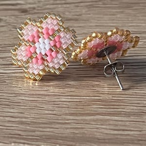 K&ouml;nnte beinhalten: Ein Paar rosa und goldene Ohrringe in Blumenform mit einem wei&szlig;en Zentrum. Die Ohrringe bestehen aus kleinen Perlen und haben einen Stiftverschluss.