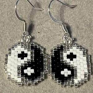 Könnte beinhalten: Handgefertigte Ohrringe mit Yin-und-Yang-Design. Die runden Ohrringe haben ein schwarz-weißes Muster und silberfarbene Haken. Die kleinen Perlen bilden eine detaillierte, strukturierte Oberfläche. Ein einzigartiges Accessoire.