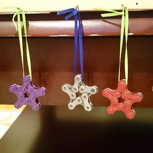 May include: Five star-shaped ornaments made from bike chains, hanging from colorful ribbons. The ornaments are blue, purple, silver, red, and gold.