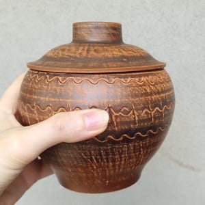 Può includere: Vaso in ceramica marrone fatto a mano con coperchio. Il vaso presenta un corpo arrotondato e un motivo ondulato decorativo. Il coperchio ha una piccola maniglia. Il vaso è tenuto in una mano.