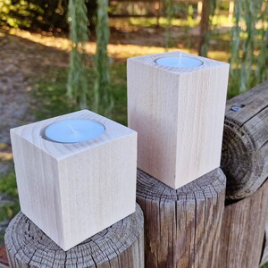 May include: Two wooden candle holders with a white tea light candle in each. The candle holders are sitting on top of wooden fence posts.