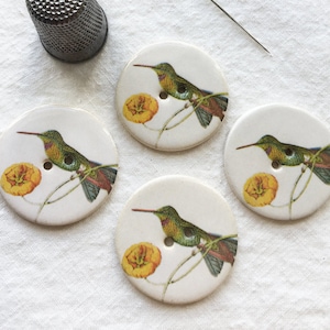 Puede incluir: Cuatro botones redondos blancos con un diseño de colibrí y flor amarilla. También hay un dedal plateado y una aguja de coser. Los botones tienen un acabado brillante y un agujero para coser.