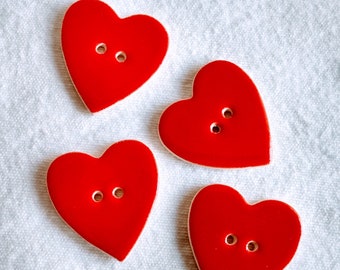 May include: Four red heart-shaped buttons with two holes each. The buttons have a glossy finish and a cream-coloured border. They are arranged on a white, textured surface.
