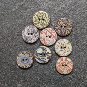 Könnte beinhalten: Eine Sammlung von acht runden Holzknöpfen mit verschiedenen floralen und gemusterten Designs. Die Knöpfe weisen eine Vielzahl von Farben auf, darunter Gelb, Rot, Blau und Braun. Jeder Knopf hat zwei Löcher zum Annähen.