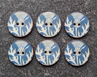 May include: A set of six circular buttons, each featuring a blue and cream-colored thistle plant illustration. Every button has two central holes for attachment, with a consistent floral pattern printed across the flat, round surface.