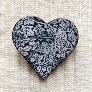 May include: Heart-shaped decorative pin with a bird and floral design in navy blue and white. The pin has a copper-colored edge and is set against a textured white background. The design features a detailed illustration of a bird amidst leaves and flowers.