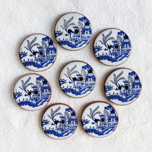 May include: Eight round buttons with a blue and white Willow pattern design. The buttons have a brown edge and two holes for sewing. The design features a bridge, buildings, and boats. The buttons are arranged on a white fabric surface.