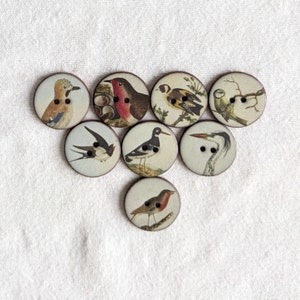 Puede incluir: Un conjunto de ocho botones redondos de madera, cada uno con una ilustración de un pájaro diferente. Los pájaros se representan en varios colores y poses, sobre un fondo crema. Cada botón tiene dos o tres agujeros para coser.