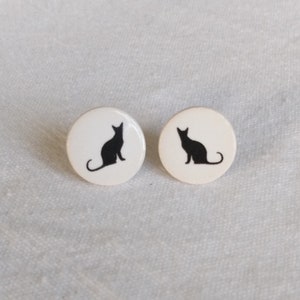Può includere: Un paio di orecchini rotondi bianchi. Ogni orecchino presenta una silhouette nera di un gatto seduto. Gli orecchini sono su uno sfondo bianco strutturato.