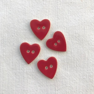 Può includere: Quattro bottoni rossi lucidi a forma di cuore, ciascuno con due fori, disposti su una superficie bianca strutturata. Questi bottoni decorativi sono ideali per cucito e artigianato.