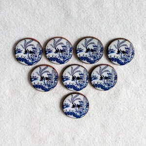 May include: Eight round buttons with a blue and white Chinoiserie pattern. Each button displays a detailed landscape scene with pagodas, boats, and bridges. The buttons have a brown edge and a black buttonhole.