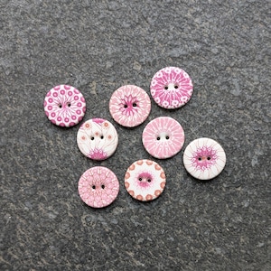 May include: A collection of eight round wooden buttons with floral patterns in shades of pink and white. Each button has two holes for sewing. The buttons vary in design, with some featuring intricate floral details.