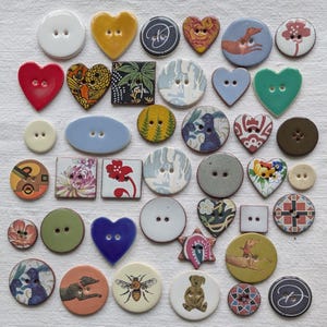 Puede incluir: Una colección de botones decorativos en varias formas, incluyendo corazones, círculos y cuadrados. Los botones presentan diversos diseños, como patrones florales, ilustraciones de animales y arte abstracto. Los colores incluyen blanco, rojo, amarillo, azul y verde.